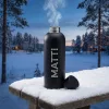 Musta personoitu termospullo höyryää lumisella pöydällä talvisessa iltahämärässä